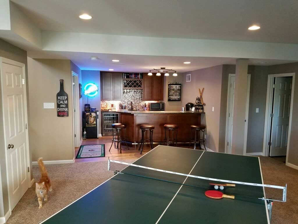 Custom basement bar setup with seating, ping pong table, and decorative signage, showcasing a cozy entertainment area ideal for gatherings and leisure in a finished basement.